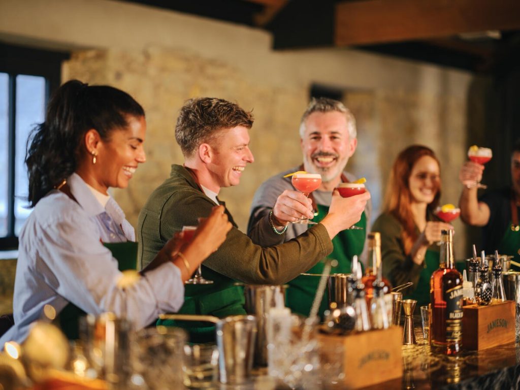 A group of people wearing green aprons raise colourful cocktails in a toast during a cocktail-making class. Bottles, bar tools and mixing equipment are arranged on the bar in front of them.