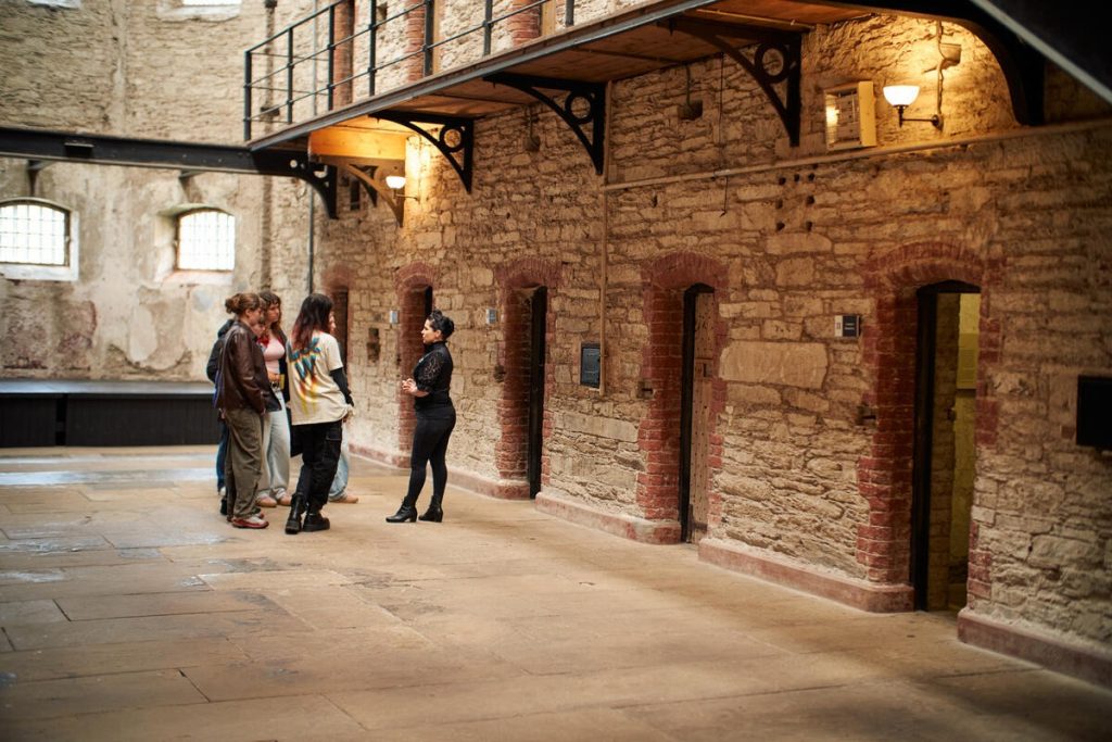 A small group stands with a guide inside a historic stone building with exposed brick walls, arched doorways and an upper-level metal walkway. Warm lighting highlights the interior's historic features.