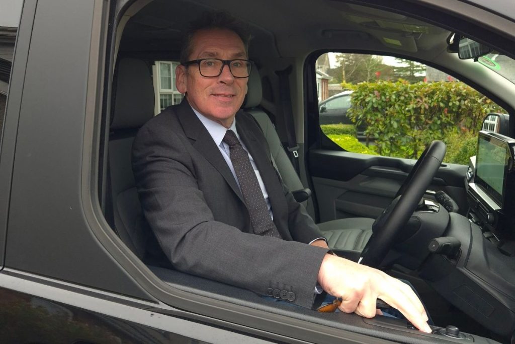 A man wearing a dark business suit and glasses sits in the driver's seat of a modern van with the window open, smiling at the camera. Houses and greenery are visible in the background.