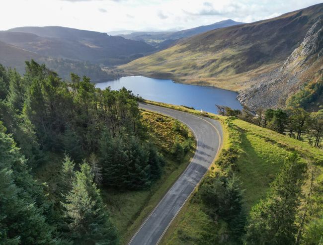 An aerial view of a winding asphalt road cutting through a lush green valley in the Wicklow Mountains, curving sharply beside a calm blue lake surrounded by forested hills and rugged, steep slopes under a cloudy sky.