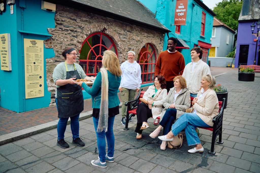 A waitress serves a tray of food to a group of smiling people gathered outside a colorful stone building with bright blue walls and red window frames.