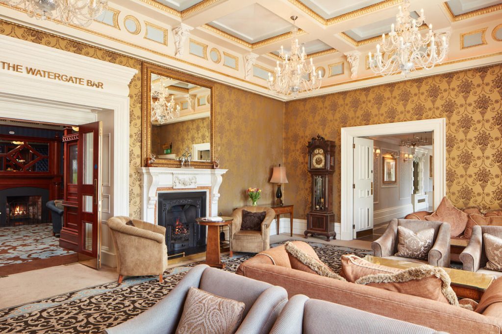 An elegant hotel lounge with patterned wallpaper, plush armchairs, a lit fireplace, ornate crystal chandeliers.